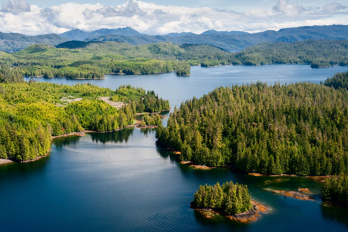 Aerial view of a winding blue lake with forested islands and distant mountains under a partly cloudy sky.